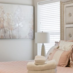 Blush pink bedroom with quilted bedspread, decorative pillows, stacked towels and a hat on the bed; beige tufted headboard and a framed tree artwork on the wall.