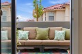 Outdoor beige sectional sofa with green and palm-pattern pillows on a patio, framed by glass doors, with a sunny backyard of beige houses and red-tiled roofs in view.