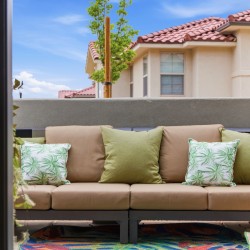 Outdoor beige sectional sofa with green and palm-pattern pillows on a patio, framed by glass doors, with a sunny backyard of beige houses and red-tiled roofs in view.