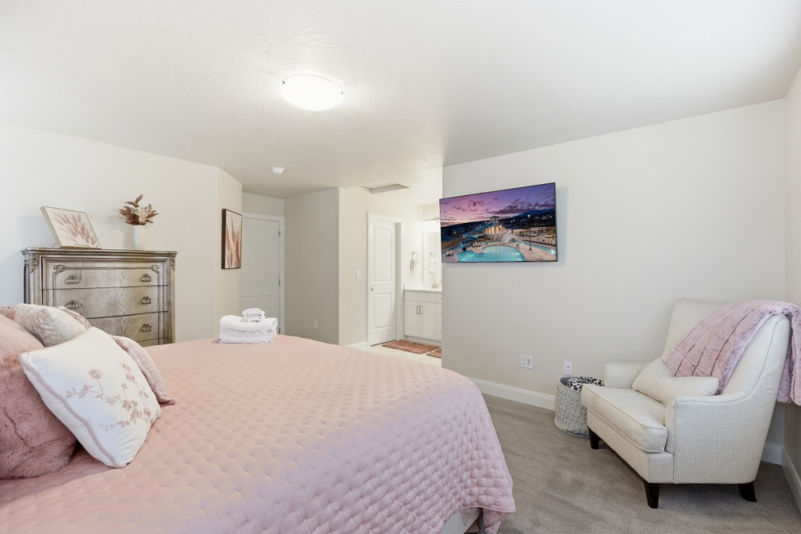 Bright bedroom with a pink quilted bed, decorative pillows, a silver dresser, and a wall-mounted TV over a chair area in the corner.