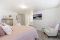 Bright bedroom with a pink quilted bed, decorative pillows, a silver dresser, and a wall-mounted TV over a chair area in the corner.