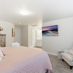 Bright bedroom with a pink quilted bed, decorative pillows, a silver dresser, and a wall-mounted TV over a chair area in the corner.