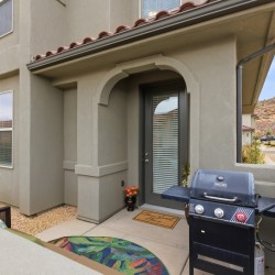 Front porch of a beige stucco townhouse with an arched entryway, glass door with blinds, and a black gas grill on the patio adjacent to the door.