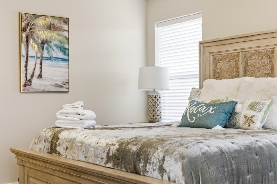 Cozy bedroom scene with a carved wooden bed, tropical-themed bedding, and decorative pillows including a teal 'Relax' pillow.
