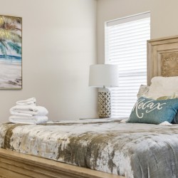 Cozy bedroom scene with a carved wooden bed, tropical-themed bedding, and decorative pillows including a teal 'Relax' pillow.
