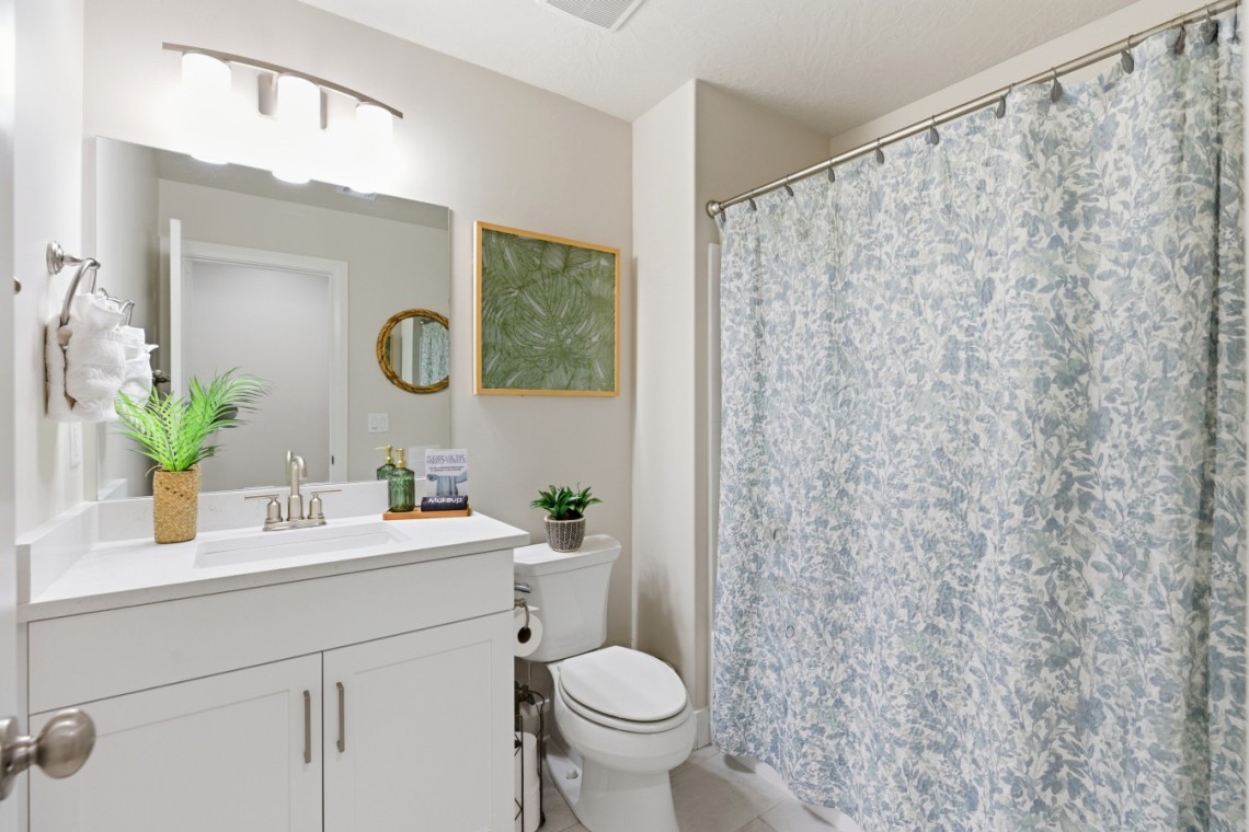 Bright bathroom with a white vanity and sink, large mirror, potted plant, green framed botanical art, and toilet beside a blue floral shower curtain.
