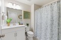 Bright bathroom with a white vanity and sink, large mirror, potted plant, green framed botanical art, and toilet beside a blue floral shower curtain.