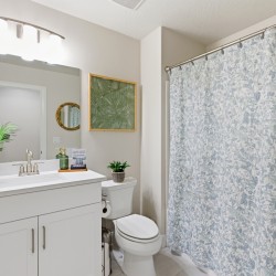 Bright bathroom with a white vanity and sink, large mirror, potted plant, green framed botanical art, and toilet beside a blue floral shower curtain.