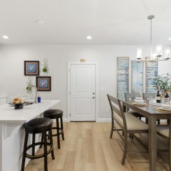 Open-concept kitchen and dining area with white cabinets, a long white island with two dark-stool seats, and a wooden table set for six under a chandelier.