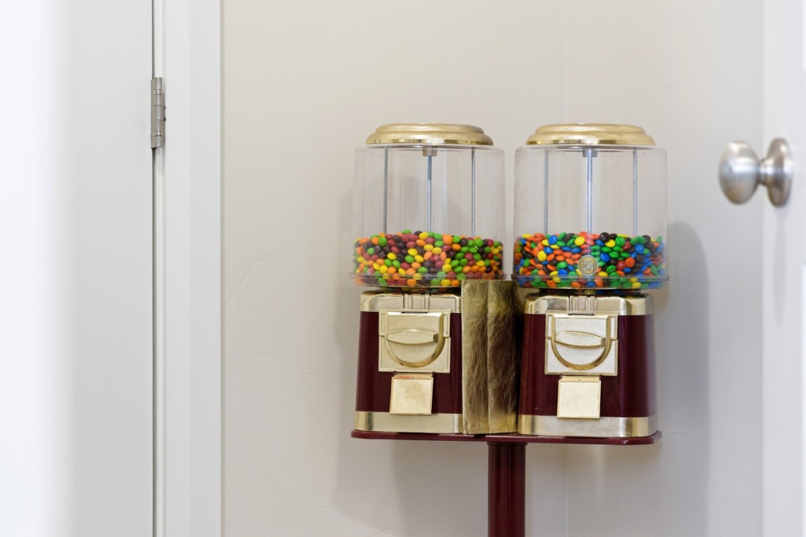 Twin old-fashioned gumball machines filled with colorful candy, mounted on a stand by a white wall and door knob nearby.