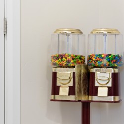 Twin old-fashioned gumball machines filled with colorful candy, mounted on a stand by a white wall and door knob nearby.