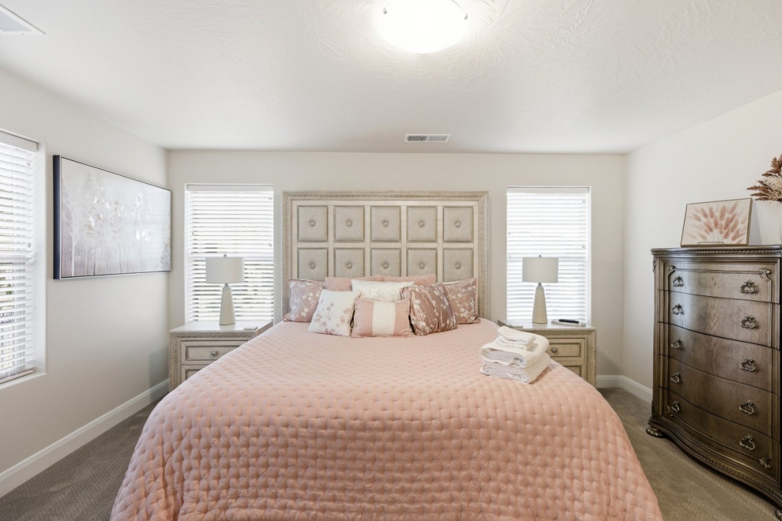 Bedroom with blush pink quilt, tufted headboard, two nightstands, and a tall ornate dresser across from the bed with lamps on each side