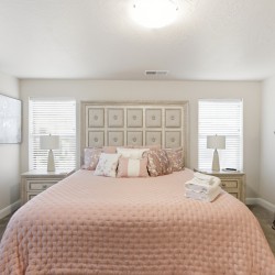 Bedroom with blush pink quilt, tufted headboard, two nightstands, and a tall ornate dresser across from the bed with lamps on each side