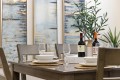 Table set for a meal with wine bottles, glasses, bowls, and woven placemats; a potted plant and blue abstract art in the background.