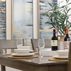 Table set for a meal with wine bottles, glasses, bowls, and woven placemats; a potted plant and blue abstract art in the background.