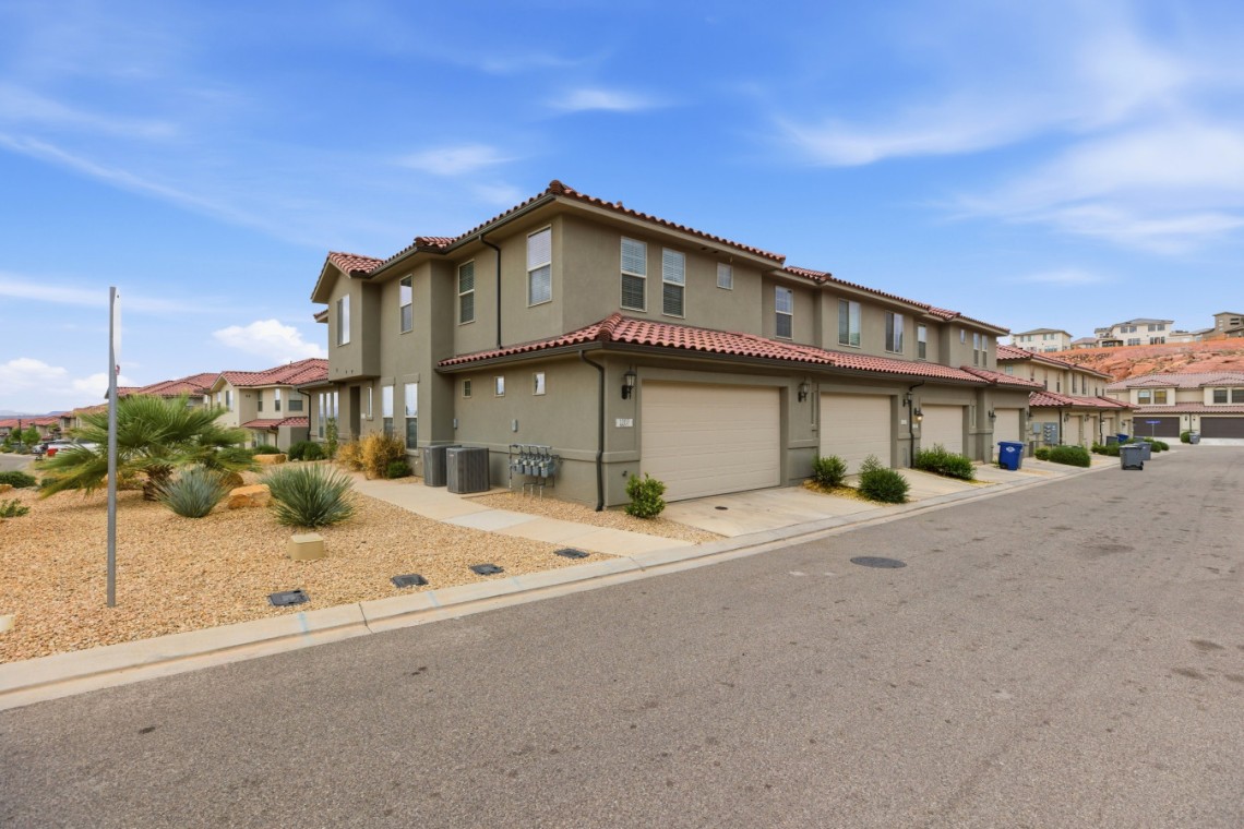Row of beige two-story townhouses with red-tile roofs and attached garages along a quiet street, desert landscaping with rocks and shrubs in the foreground under a blue sky.