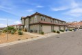 Row of beige two-story townhouses with red-tile roofs and attached garages along a quiet street, desert landscaping with rocks and shrubs in the foreground under a blue sky.
