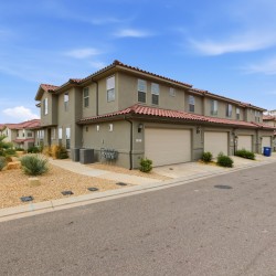 Row of beige two-story townhouses with red-tile roofs and attached garages along a quiet street, desert landscaping with rocks and shrubs in the foreground under a blue sky.