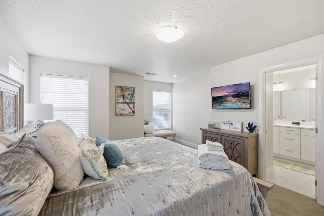 Bright master bedroom with a tufted gray bed, plush pillows, and folded towels on a textured duvet; soft natural light from two windows. A wall art palm tree and a bench sit near the windows. A dresser with a TV, decorative sign, and blue coral decor sits opposite the bed; bathroom visible through open door.
