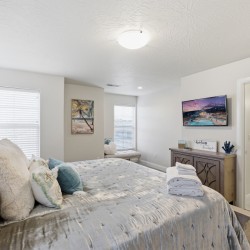 Bright master bedroom with a tufted gray bed, plush pillows, and folded towels on a textured duvet; soft natural light from two windows. A wall art palm tree and a bench sit near the windows. A dresser with a TV, decorative sign, and blue coral decor sits opposite the bed; bathroom visible through open door.