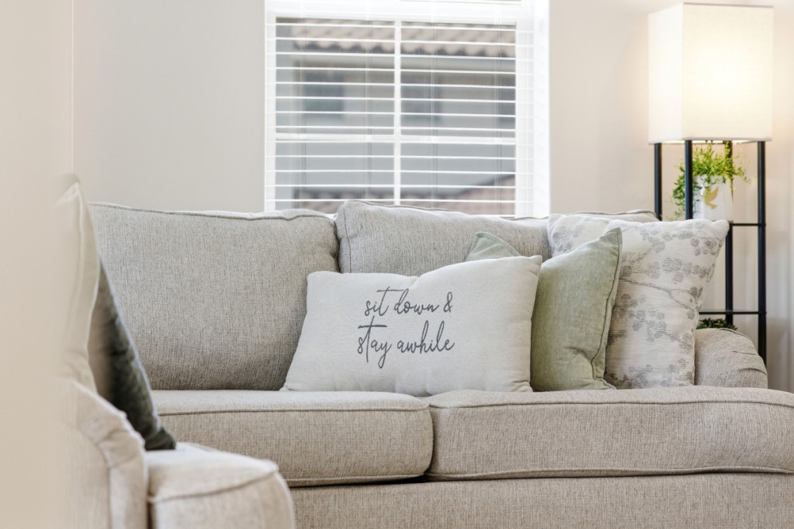 Neutral-toned living room with a beige/gray upholstered sofa and multiple decorative pillows, including a white pillow that reads 'sit down & stay awhile'.