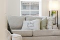 Neutral-toned living room with a beige/gray upholstered sofa and multiple decorative pillows, including a white pillow that reads 'sit down & stay awhile'.