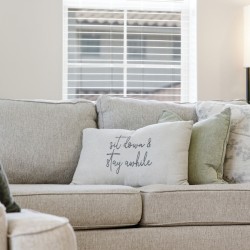 Neutral-toned living room with a beige/gray upholstered sofa and multiple decorative pillows, including a white pillow that reads 'sit down & stay awhile'.