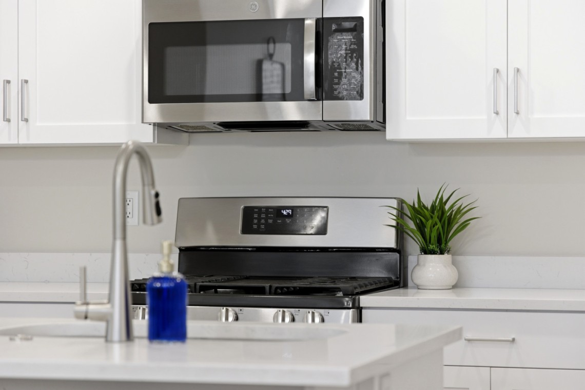 Modern kitchen scene with stainless steel microwave above a gas range, white cabinets, and a small potted plant on the counter.