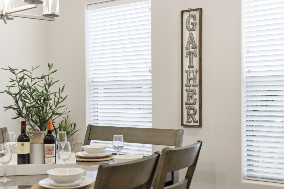 Dining room table set for a meal with bowls, glasses, wine bottles, and a potted plant nearby; a rustic vertical GATHER sign on the wall.
