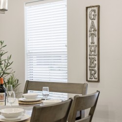 Dining room table set for a meal with bowls, glasses, wine bottles, and a potted plant nearby; a rustic vertical GATHER sign on the wall.