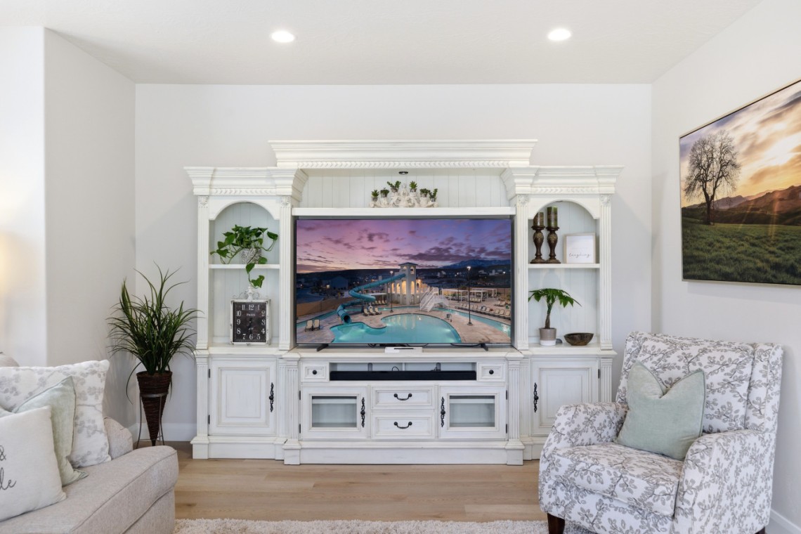 Elegant white entertainment center surrounding a flat-screen TV showing a poolside resort scene, with plants and decor on the shelves around it.