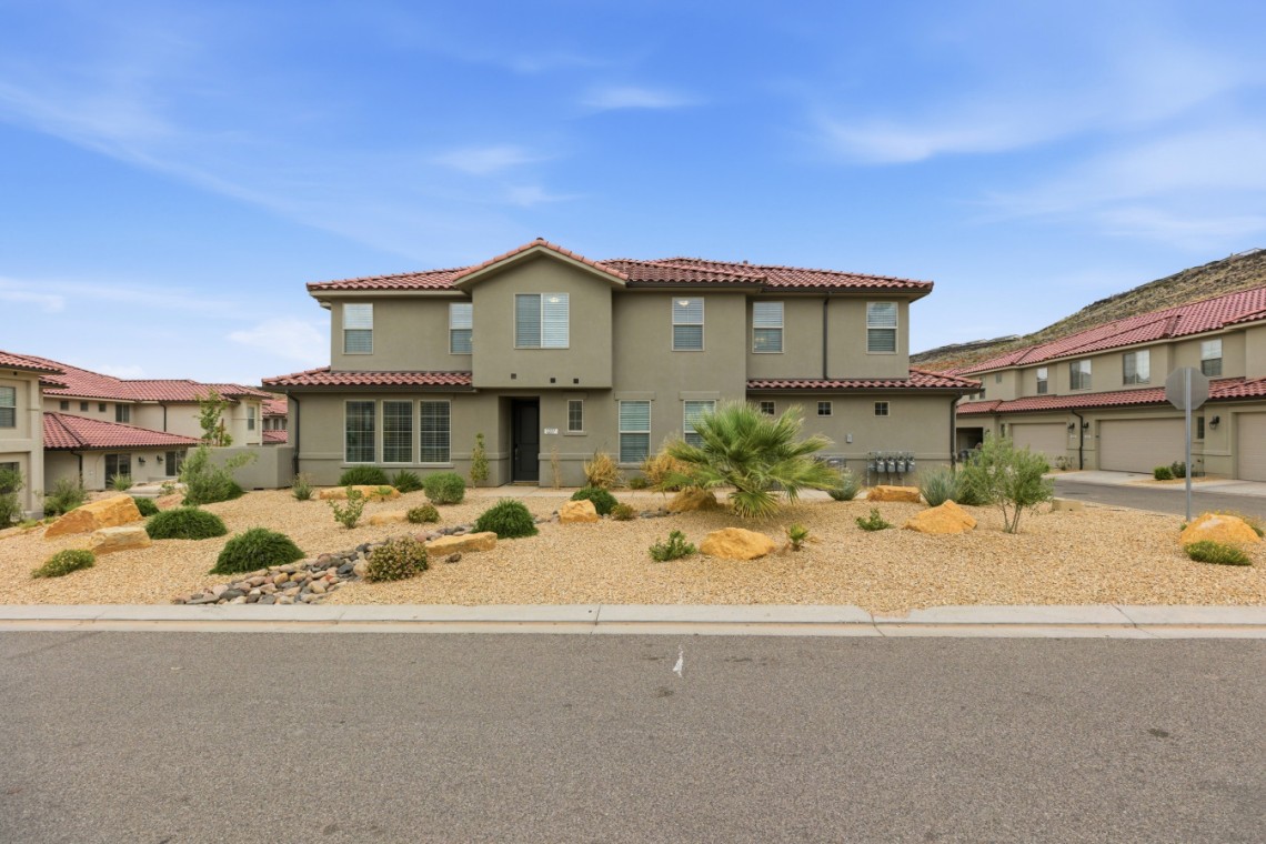 Two-story beige stucco house with a red tile roof and a desert rock yard in front of a suburban street