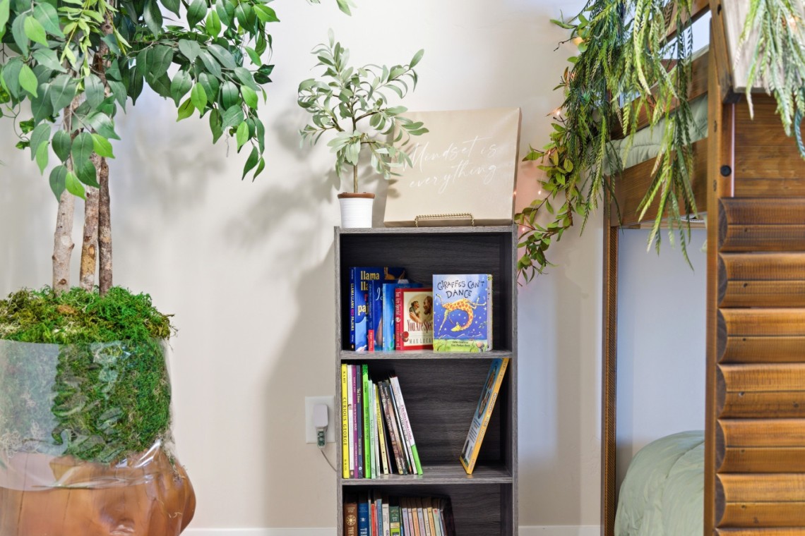 Gray wooden bookshelf framed by lush green plants, with a beige sign reading 'Mindset is everything' on top.