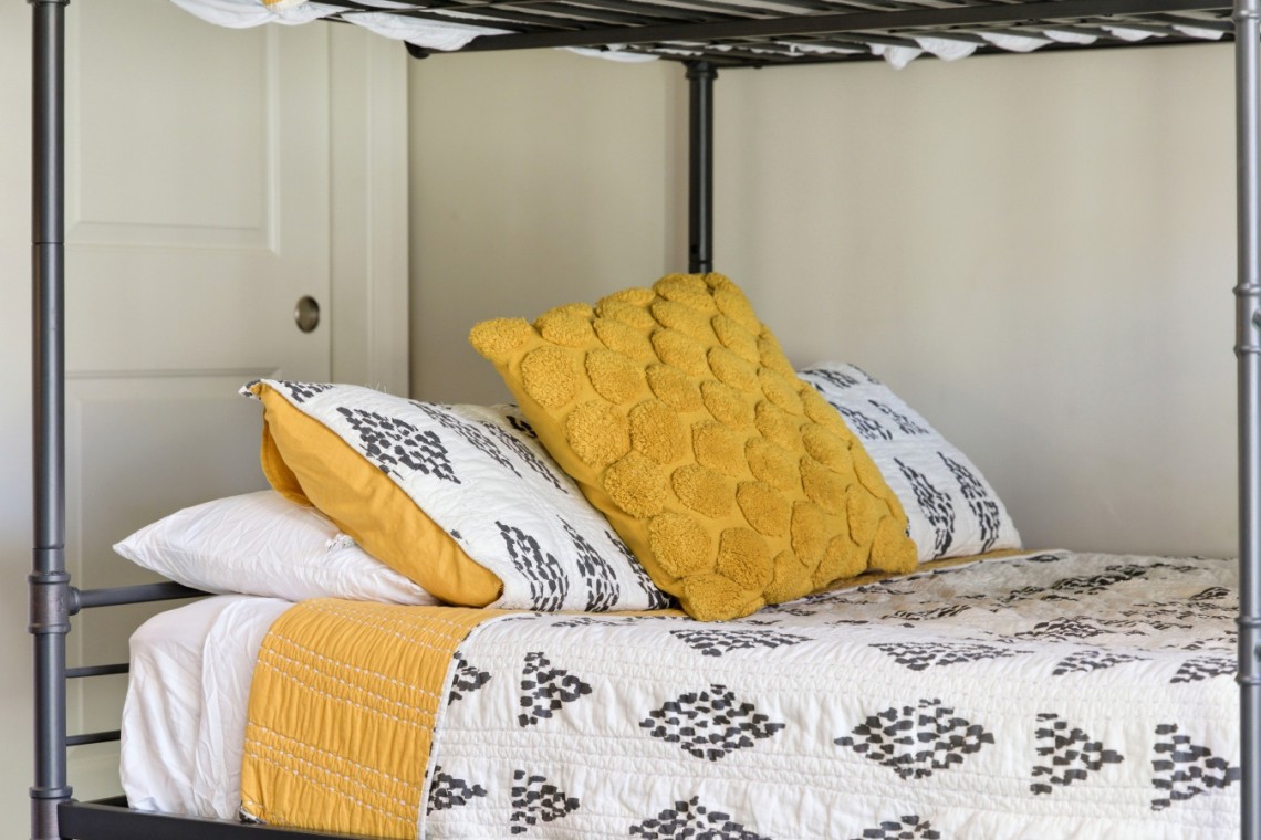 A neatly made bed with a white quilt featuring black geometric patterns and yellow accent pillows against a metal frame bed.