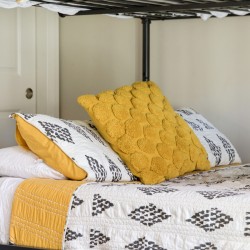 A neatly made bed with a white quilt featuring black geometric patterns and yellow accent pillows against a metal frame bed.