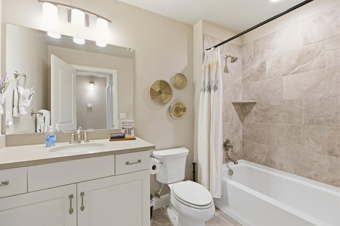 Bright bathroom with a white vanity and sink, large mirror, three woven baskets on the wall, a toilet, and a tiled tub/shower with a white curtain.
