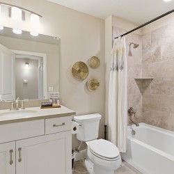 Bright bathroom with a white vanity and sink, large mirror, three woven baskets on the wall, a toilet, and a tiled tub/shower with a white curtain.