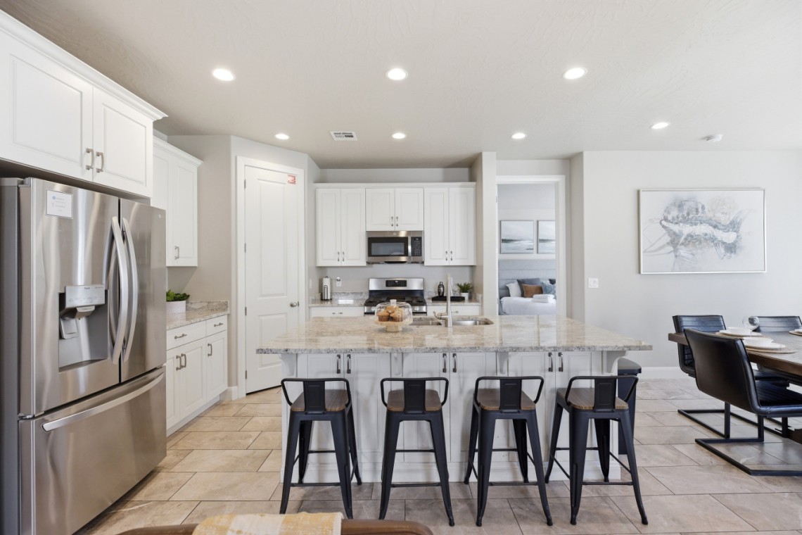 Open-concept kitchen with white cabinets, stainless steel fridge, granite island and six black bar stools at the counter.