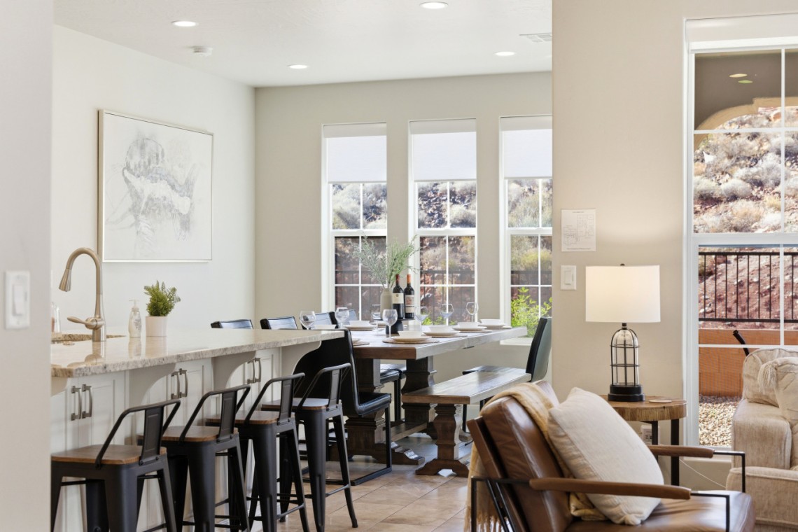 Open-concept kitchen and dining area with a marble island, black metal stools, and a wooden dining table set for dinner by bright windows.