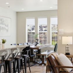 Open-concept kitchen and dining area with a marble island, black metal stools, and a wooden dining table set for dinner by bright windows.