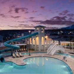 Community pool with a teal water slide, beige clubhouse, and lounge chairs at sunset with mountains in the distance.
