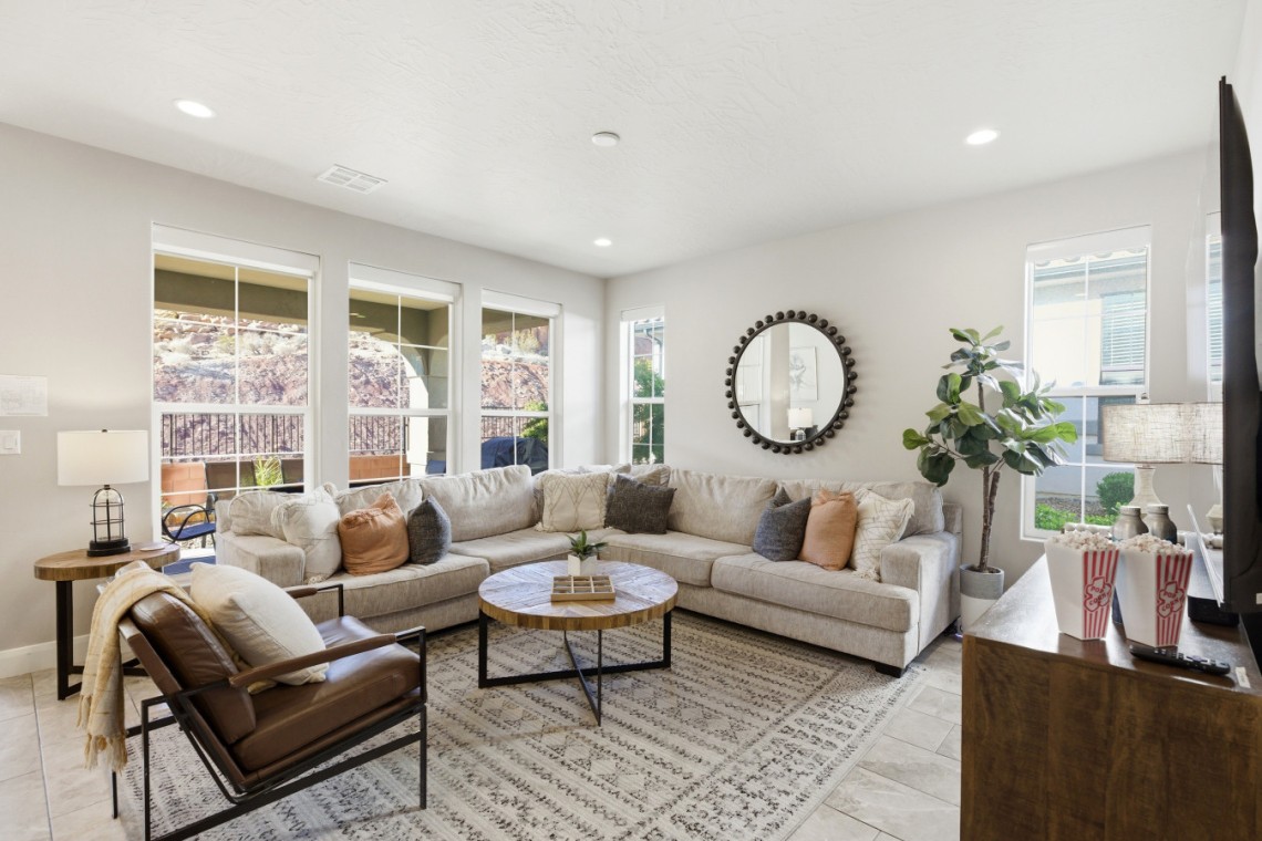 Bright living room with a beige sectional, brown accent chair, round wooden coffee table, and a textured gray rug under neutral decor pieces.
