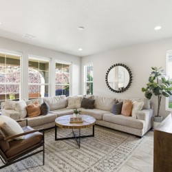 Bright living room with a beige sectional, brown accent chair, round wooden coffee table, and a textured gray rug under neutral decor pieces.