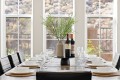 Dining table set for a meal with white bowls and wine glasses, a central vase with greenery, and two bottles of wine. Large windows in the background.
