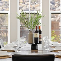 Dining table set for a meal with white bowls and wine glasses, a central vase with greenery, and two bottles of wine. Large windows in the background.