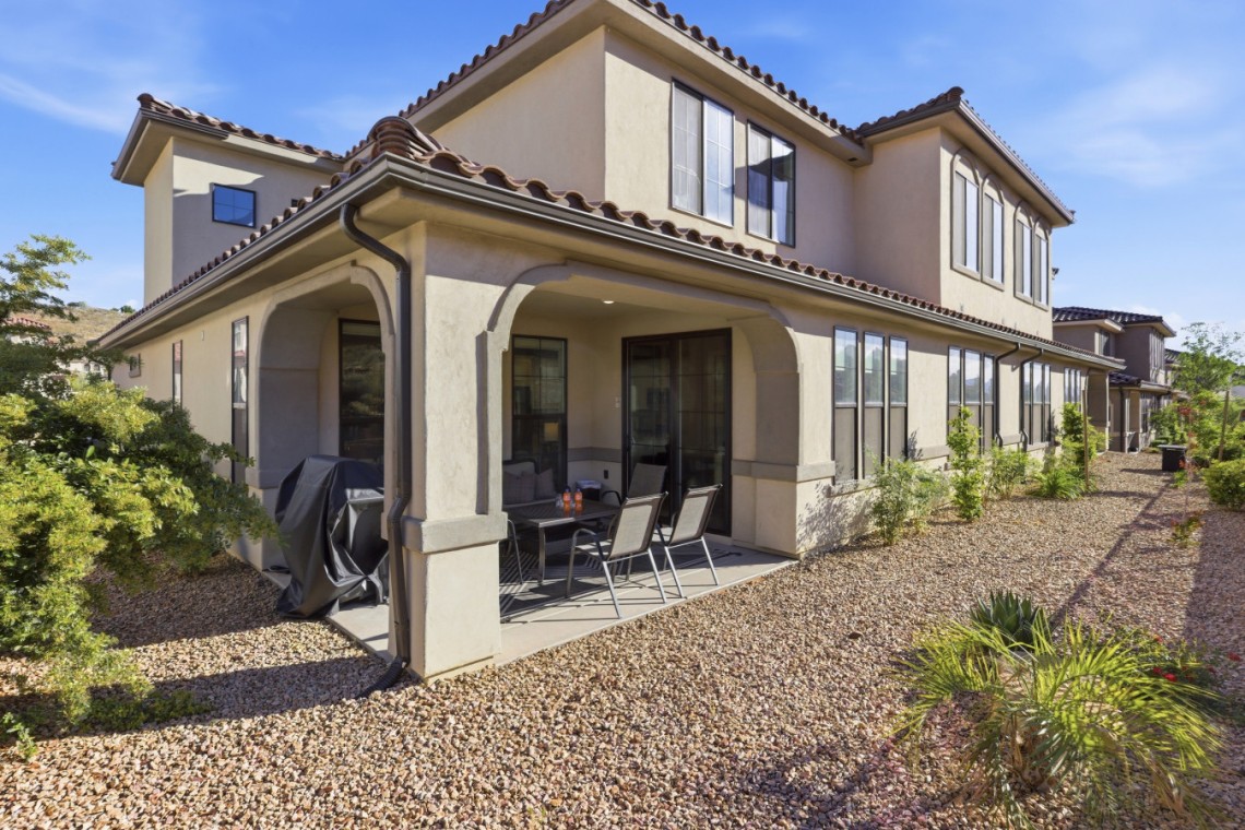 Row of beige stucco townhouses with tile roofs; covered patio, outdoor table and chairs, and gravel landscaping under a blue sky.