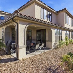 Row of beige stucco townhouses with tile roofs; covered patio, outdoor table and chairs, and gravel landscaping under a blue sky.