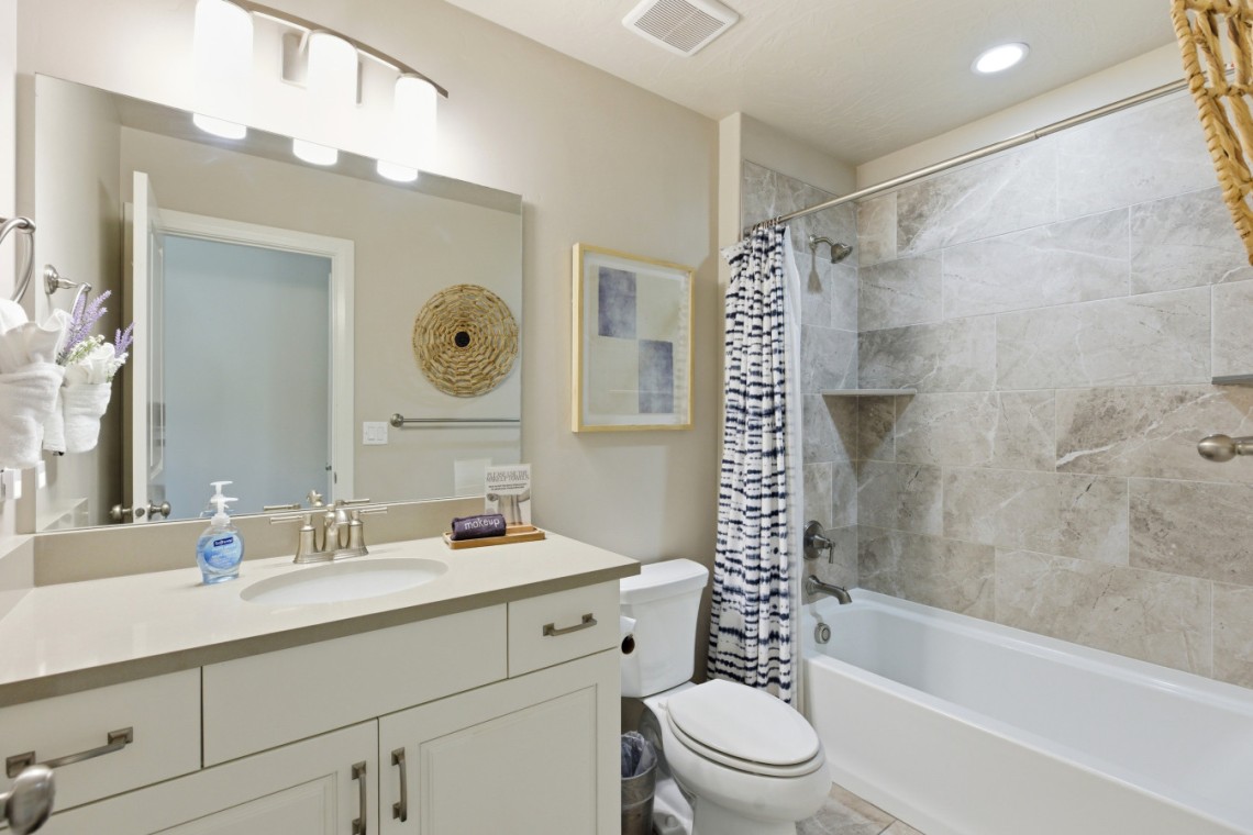 Bright bathroom with a white vanity, large mirror, and a striped shower curtain beside a tiled tub area.