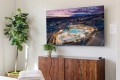 Large wall-mounted TV shows an aerial view of a resort pool at sunset; a wooden cabinet and two potted plants sit below in a modern living room.