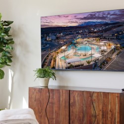 Large wall-mounted TV shows an aerial view of a resort pool at sunset; a wooden cabinet and two potted plants sit below in a modern living room.